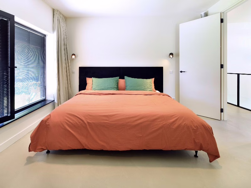 Naadloze gietvloer in een rustige slaapkamer met tweepersoonsbed en terracotta beddengoed